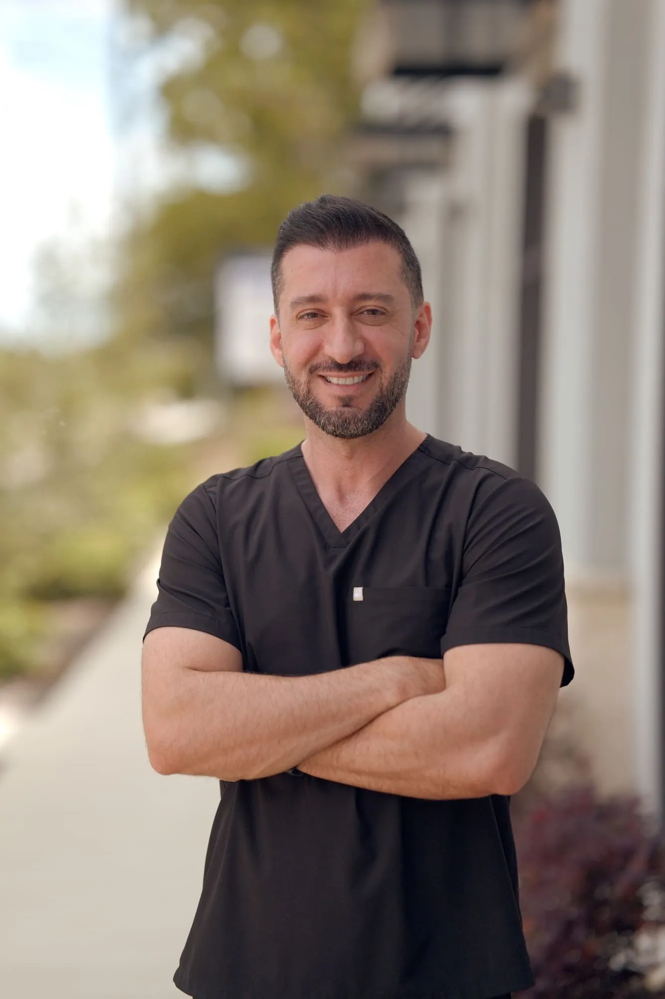 Outdoor professional portrait of a smiling man wearing black medical scrubs with his arms crossed