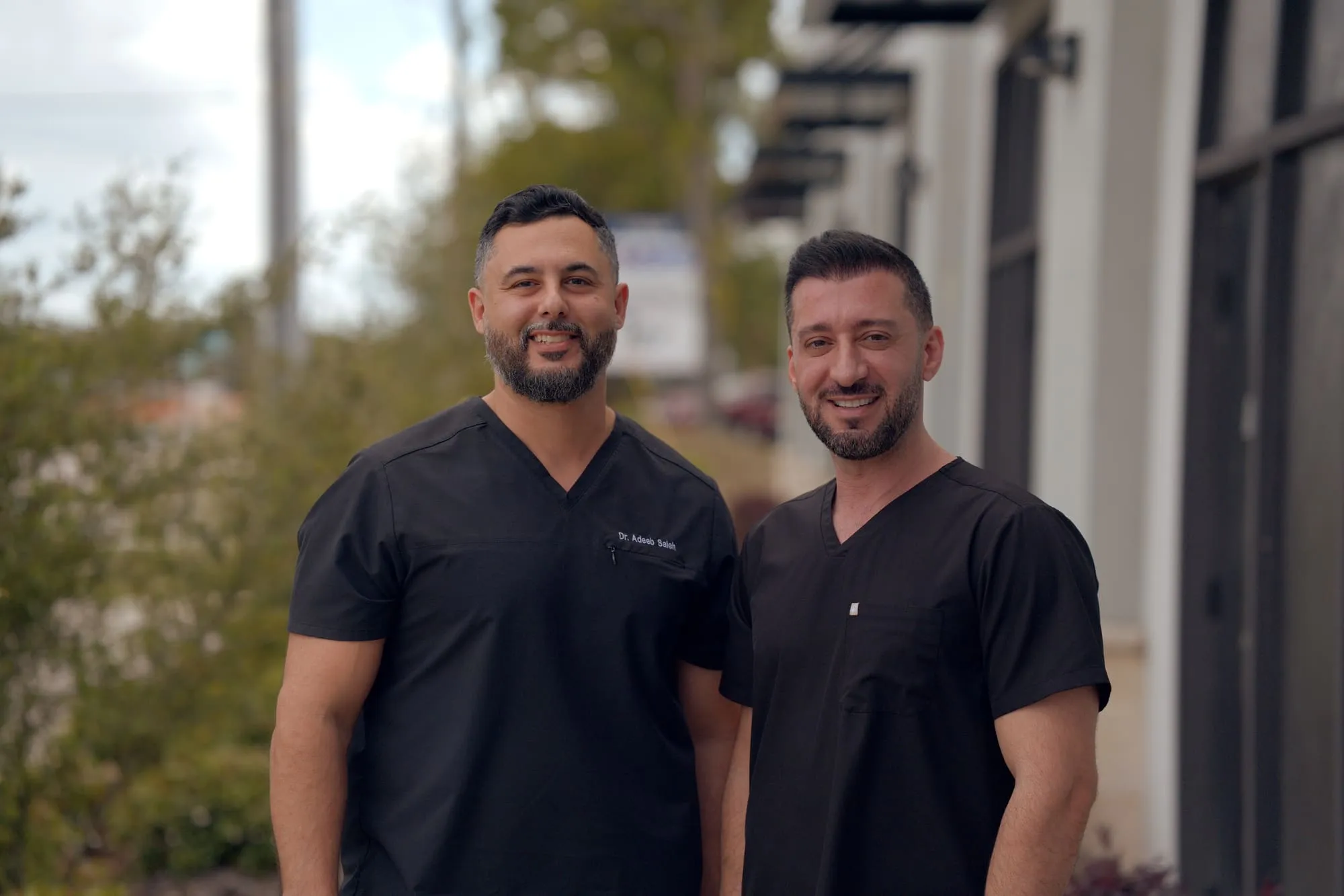 Two smiling medical professionals standing together outdoors in black scrubs
