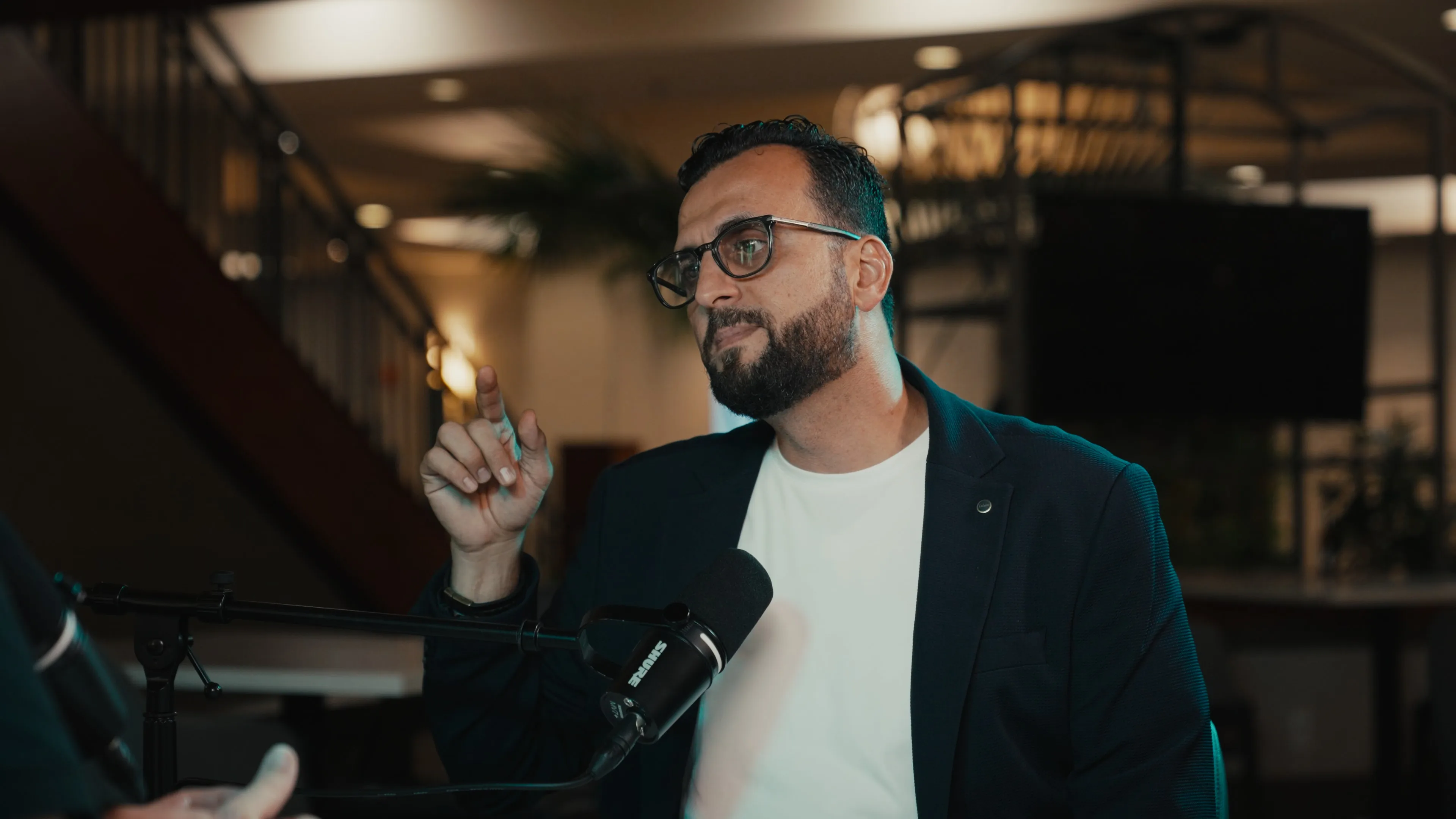 Ya Hala host Haithum speaking into a Shure MV7+ microphone with warm hotel lights and a teal edge light behind him