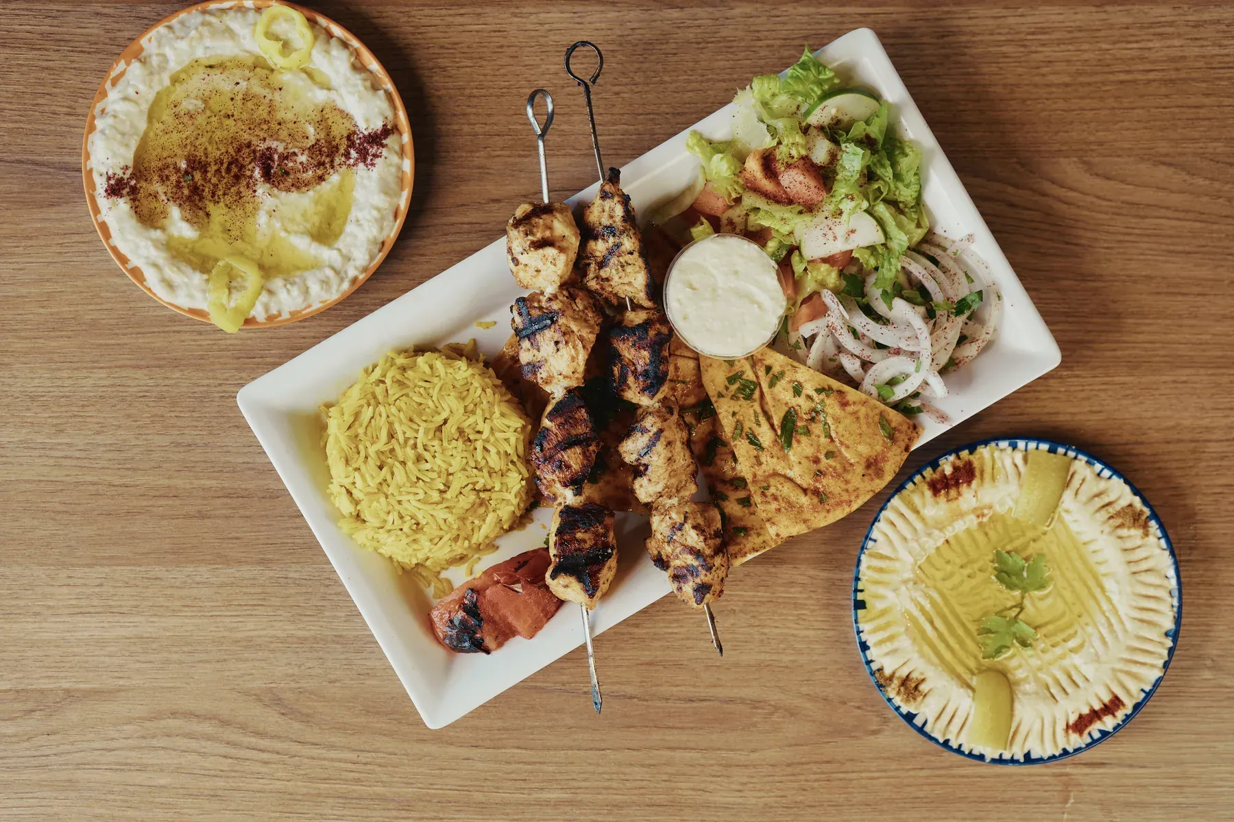 Grilled chicken skewers with rice, salad, pita bread, hummus, and baba ghanoush photographed from above