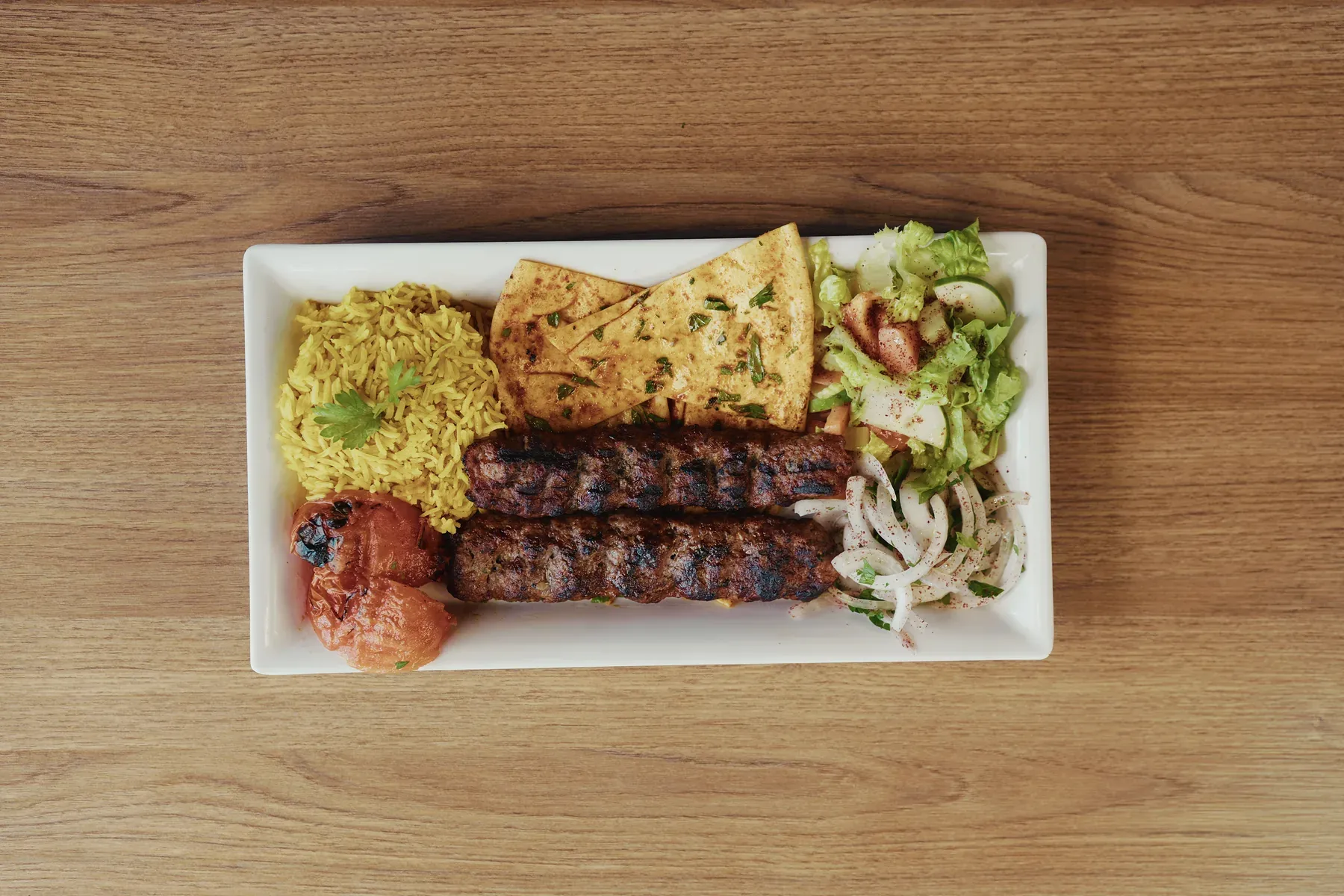 Two grilled kafta kebabs served with rice, salad, onions, tomato, and seasoned pita bread