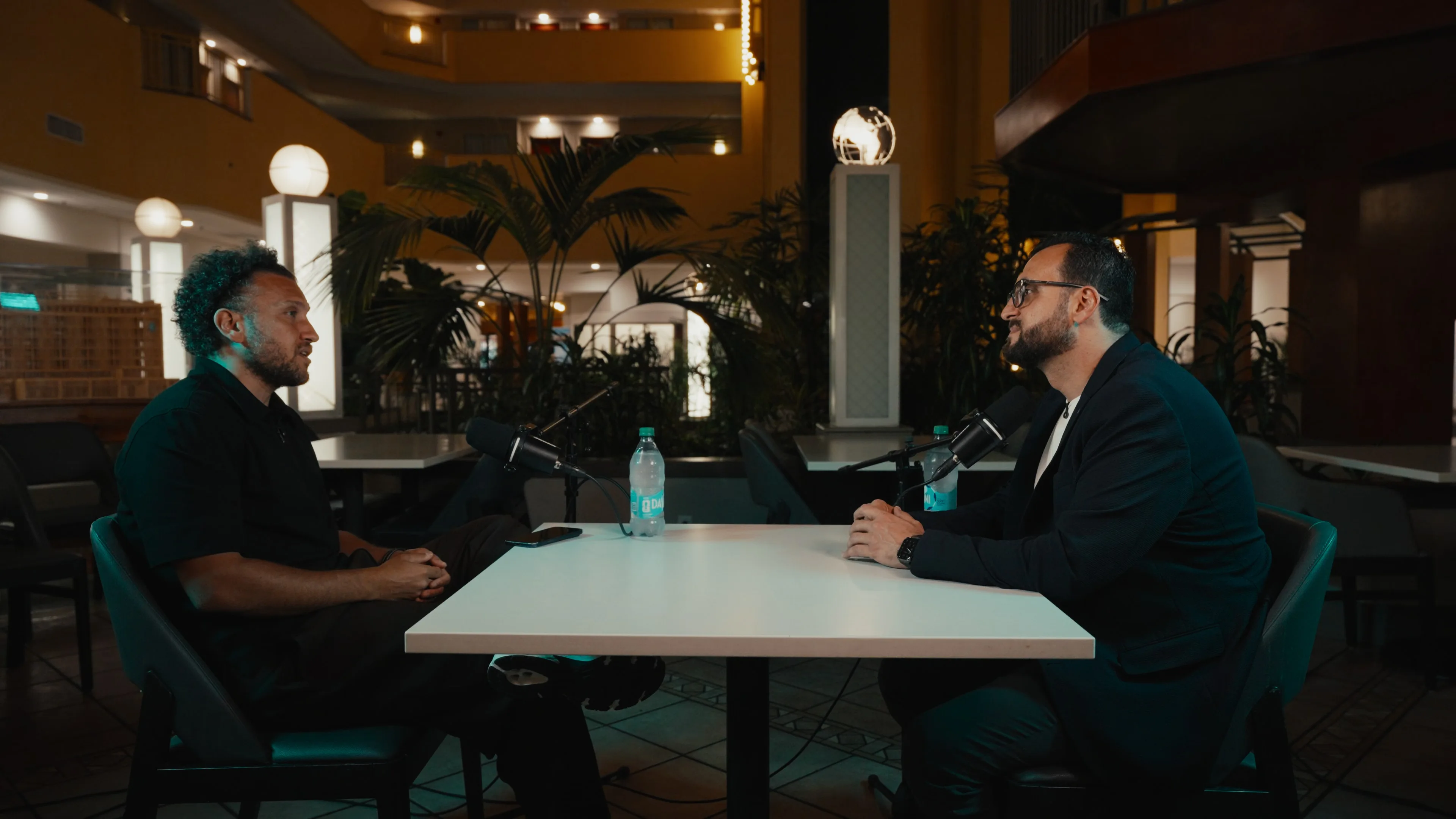 Haithum interviewing Egyptian comedian Youssef Hussein at a table in the Embassy Suites Tampa USF atrium, lit with soft key light and teal edge light